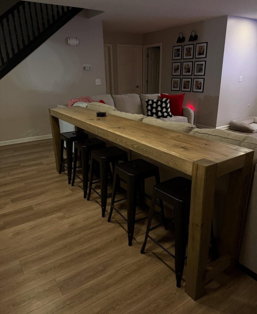 Wooden bar table with black stools positioned behind a grey sectional sofa.